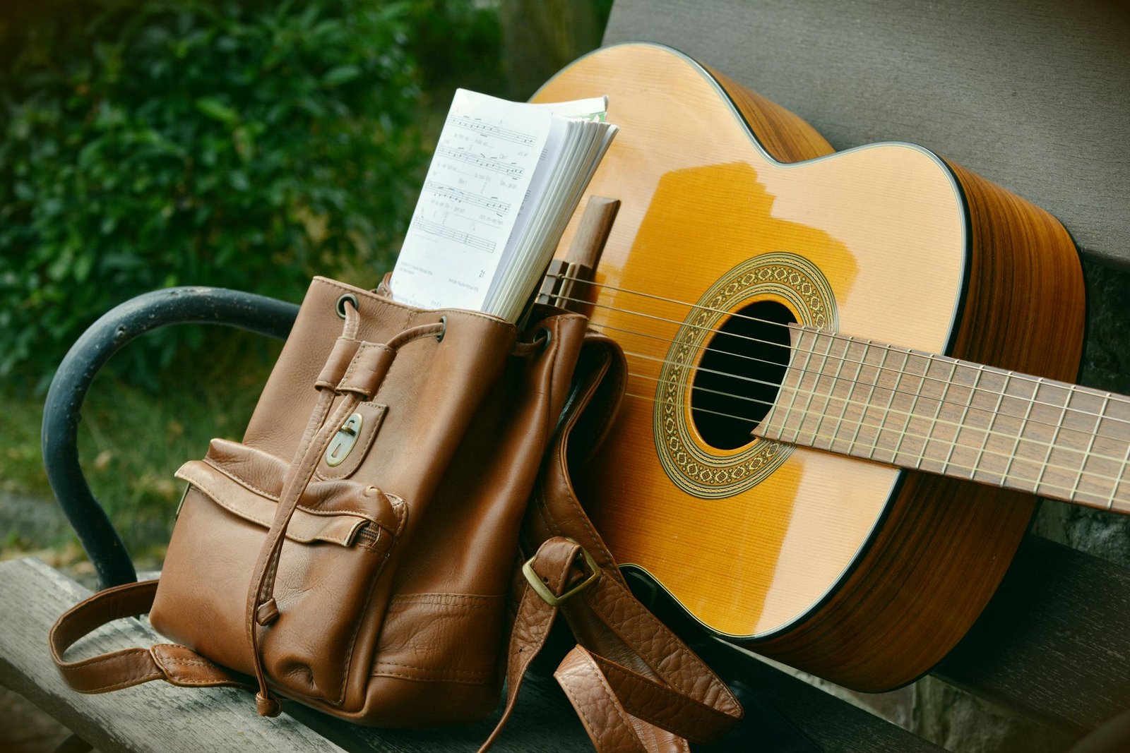 A serene outdoor scene of an acoustic guitar and a brown backpack on a wooden bench, perfect for music lovers.